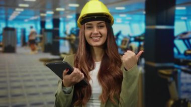 Spor salonundaki tabletle gülümseyen kadın başparmağıyla onaylıyor. Modern bir spor merkezinde başarılı olduğunu gösteriyor..