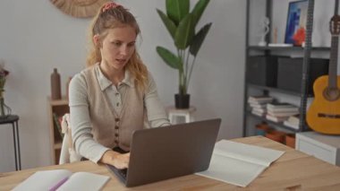 Evde dizüstü bilgisayarı olan bir kadın başını genç sarışın kilitlerin arkasına eğiyor ve modern bir iç mekanda masa başında oturuyor..