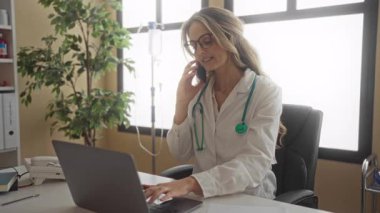 Steteskoplu kadın doktor, klinik odasındaki iç mekanda bilgisayarla telefonla konuşuyor..