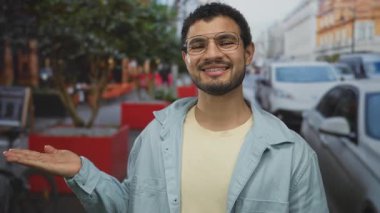 Şehrin hareketli caddesinde park etmiş arabaları ve kırmızı saksılarıyla açık el hareketleriyle İspanyol genç adam iyimserlik.