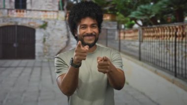 Gülümseyen adam bir şehir caddesinde, ağaçlar ve kaldırımlarla kentsel bir ortamda pozitiflik ve bağlılık taşıyarak neşeyle işaret ediyor..