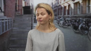 Sarışın kadın park halindeki bisiklet ve tuğla binalarla dolu bir caddede yüzünü gösteriyor..