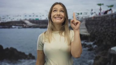 Sahilde gülümseyen bir kadın sahilde arka planda beyaz binaları işaret ederek bulutlu bir gökyüzünün altında açık denizde neşeli bir ifade sergiliyor..