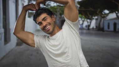 Güneş ışığıyla aydınlanmış kaldırım taşı caddesinde kalp oluşturan silahlı bir adam gülümsüyor ve kameraya bakıyor..