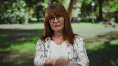 Gözlüklü kıdemli İspanyol kadın parkta duruyor ve açık havada canlı yeşil bir arka plana karşı x şeklinde kollarını kavuşturuyor..