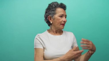 Beyaz gömlekli, kırmızı bilezik takan, gösterişli bir ifade ve zarafeti olan, gri saçlı yaşlı kadın, canlı mavi arka planda ellerini yana uzatıyor..