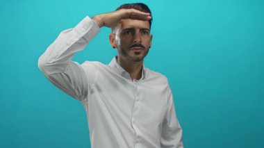 Stüdyoda turkuaz duvara karşı beyaz gömlek giymiş, uzaktan bakan gözleri koruyan bir adam..