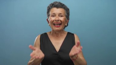 Mavi bir arka planda mutluluğu ifade eden gri saçlı yaşlı bir kadın, canlı bir gülümseme ve açık ellerle siyah giyinmiş, neşe ve pozitiflik taşıyor..