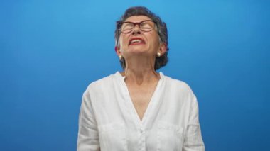 Duygularını kapalı gözlerle ifade eden ve saf mavi arkaplan karşısında beyaz bluz ve gözlük takan bir kadın..