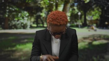 Açık havada güneşli, yemyeşil bir parkta gülümseyen ve kamerayı işaret eden genç bir kadın..