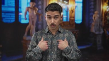 Müzedeki genç İspanyol adam, arka planda heykeller var. Kendini işaret ediyor ve şaşırtıyor ya da sorguluyor..