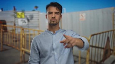 Yüzünde ciddi bir ifade olan genç bir adam açık hava inşaatında jestleri durduruyor..