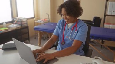 Kadın doktor, dizüstü bilgisayarında boynunda kırmızı steteskop ve arkasında da muayene masası olan bir binanın içinde yazıyor..