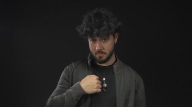 Sakallı ve kıvırcık saçlı genç adam siyah bir arkaplana karşı jest yapıyor, koyu renk ceketli ve gündelik kıyafetli oyuncu ve düşünceli bir tavır sergiliyor..