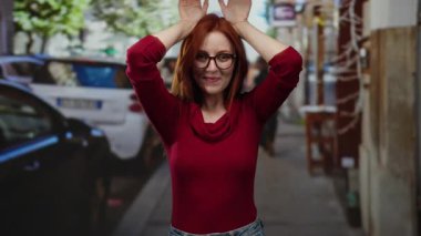 Gözlüklü ve kızıl saçlı bir kadın, neşeli bir şekilde gülümsüyor, elleri başının üstünde, hareketli bir caddede kulak simülasyonu yapıyor..