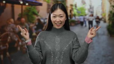Şehirli bir terasta gülümseyen ve parmakla işaret eden bir kadın hareketli bir şehir sokak ortamında gri bir süveter giyip açık hava ortamı sergiliyor..