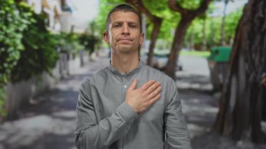 Sokakta dikilen bir adam bir eli göğsünde, diğeri yeşil ağaçların arasında yükselmiş bağlılık ve samimiyetini gösteren bir jest yapıyor..