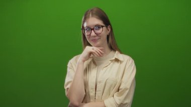 Gözlüklü genç sarışın kadın, stüdyoda yeşil perde arkası gömlekli, ince gülümsemeli çeneye dokunur..