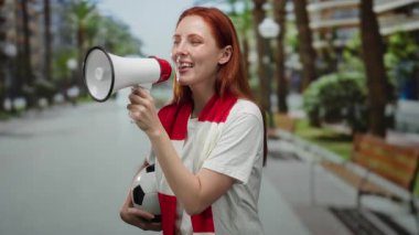 Şehir caddesinde kırmızı eşarp takmış megafonla ve futbol topuyla tezahürat yapan bir kadın.