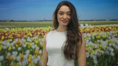 Yeşil bir çiçek parkında duran kadın, saç eserken canlı lale çiçeklerini işaret ediyor; bahar neşesi.