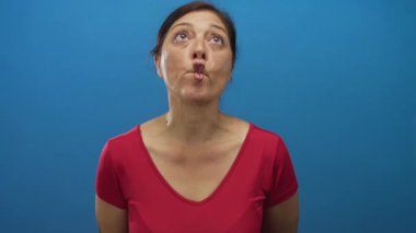 Orta yaşlı, siyah saçlı, kırmızı gömlekli, dudak çantalı ve stüdyoda ileriye bakan bir kadın..