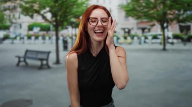 Ağaçlarla dolu bir caddede gülümseyen bir kadın, kızıl saçlı ve gözlüklü, şehir ortamında güzelliği ve zarafeti somutlaştıran bir kadın..