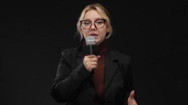 Siyah arka plan duvarına mikrofon dayamış gözlüklü kadın muhabir profesyonel ve kendinden emin haber yayını yapıyor..