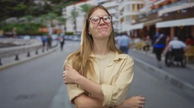 Sarışın, gözlüklü bir kadın, güneşli bir şehir caddesinde kollarını kavuşturuyor. Kafeler ve bulanık yayalarla dolu. Kendine güveni tam..