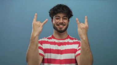 Sakallı genç adam gülümsüyor ve mavi arka plana taş hareketi yapıyor. Gözlük ve çizgili kırmızı gömlek giyiyor. Mutluluk ve enerji taşıyor.