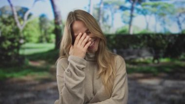 Sarı saçlı, bej renkli kazak giyen bir kadın gülümseyip, yeşil parkta eliyle yüzüne dokunuyor; neşe kahkahaları sıcaklığı.