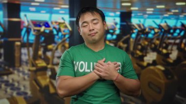 Yeşil gönüllü tişörtü giyen genç adam arka planda egzersiz ekipmanları, somut kuvvet ve toplum ruhuyla modern spor salonunda güven içinde duruyor..