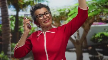 Gri saçlı, kırmızı elbiseli son sınıf öğrencisi, neşeli bir şekilde dans ediyor. Canlı yeşil bir parkta, gür ağaçlar ve bitkilerle çevrili, akıllı telefonuyla müzik dinliyor..