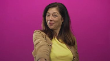 Sarı gömlekli kadın pembe stüdyoda kameraya doğru elini uzatıyor; güven bağlantısını selamlıyor.