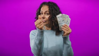 Orta yaşlı bir kadın, canlı bir mor arka plana karşı Peru banknotları tutuyor, şakacı bir şekilde para ve sevgiyi simgeleyen bir el hareketi yapıyor..