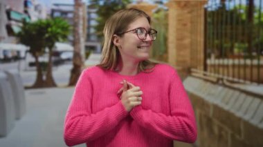 Pembe kazaklı, gözlüklü, gülümseyen ve parmağıyla şehir caddesini işaret eden, arkasında tuğla bina duvarı olan bir kadın..