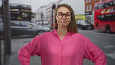 Pembe tişörtlü kadın şehir caddesinde neşeli bir ifadeyle duruyor. Etrafı arabalarla çevrili ve şehir planıyla dolu..