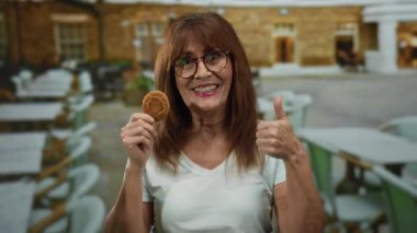 Sokak terasında gülümseyen son sınıf İspanyol kadın. Elinde kurabiyeyle bir açık hava restoranında jest yapıyor..