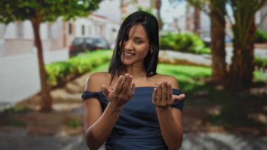 Güneşli bir parkta el kol hareketi yapan genç bir kadın, arka planda ağaçlarla ve şehir sokağıyla güvenle gülümsüyor, açık bir şehir ortamında sıcaklık ve davetiye taşıyor..