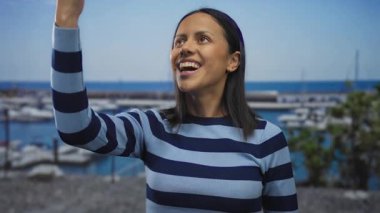 Güneşli bir günde deniz kenarında gülümseyen bir kadın çizgili bir gömlek giyiyor, etrafı tekneler ve canlı mavi suyla çevrili, neşeli, rahat bir yaz havası sergiliyor..