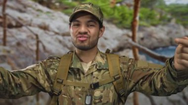 Kamuflaj üniformalı adam, görünür künyeli kayalık sahil plajının yanında kendi göğsünü işaret ediyor..