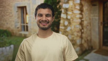 Adam taş bina girişinin önünde serçe parmağı ve baş parmağıyla telefon hareketi yapıyor; dostça iletişim.