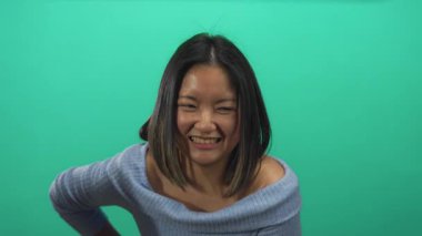 Mavi kazaklı kadın, stüdyo ortamında tek başına kalırken karın ağrısı ya da rahatsızlık hissi veren yeşil bir zemine karşı huzursuzluk içinde..