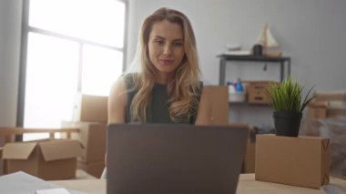 Genç bir kadın yeni oturma odasında yeni bir dizüstü bilgisayara bakıyor. Kutuları ve bitkileri taşıyor. Yeni bir başlangıç ya da ofis kurulumu öneriyor..