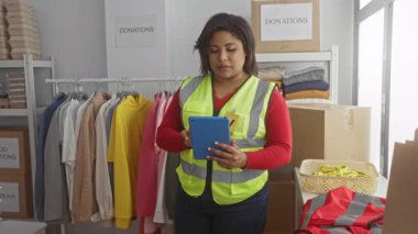 Yansıtıcı yelek giyen gönüllü bir kadın bağış merkezinde tablet kullanıyor. Kıyafetler ve kutularla çevrili..