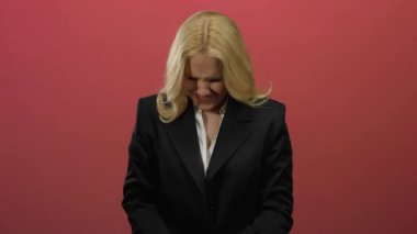 Sarı saçlı kadın, canlı kırmızı bir arkaplan önünde sıkıntılı görünüyor. Resmi siyah takım elbisesiyle duygularını ve gerilimini ifade ediyor..