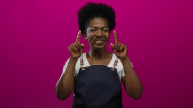 Önlüklü genç kadın gülümsüyor ve doğrudan kameraya bakıyor. İzole edilmiş pembe arka planda, kendine güven ve yakınlık yayıyor.