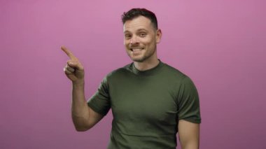 Gülümseyen ve coşkuyla işaret eden genç bir adam canlı pembe bir arka planda izole edilmiş bir stüdyoda neşeli ifadeler ve kendinden emin tavırları ortaya koyuyor..
