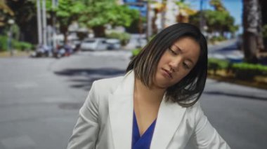 Güneşli bir caddede beyaz ceketli bir kadın modern mimari ve yeşil ağaçları sergileyen kentsel bir arka plana karşı düşünceli görünüyor.