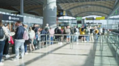 Kalabalık yoğun bir havaalanı terminalinde bekliyor. Geçmişi bulanık ve yoğun bir atmosfer yayıyor..