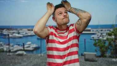 Çizgili tişörtlü genç adam deniz kıyısında tekneleri ve açık mavi gökyüzü arkaplanıyla şakacı hareketler yapıyor.